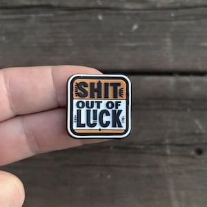 May include: A black and white enamel pin with an orange border. The pin says "SHIT OUT OF LUCK" in a bold, black font.