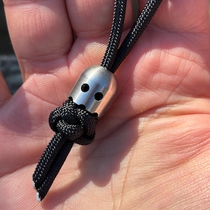 Op de afbeelding: Close-up van een zwart koord met een zilverkleurige metalen kraal. De kraal heeft twee ronde gaten en is met een knoop aan het koord bevestigd. Het koord wordt in de hand van een persoon gehouden.