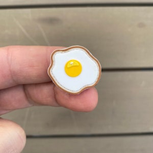 May include: A shoe charm in the shape of a fried egg. The charm has a white egg white, a yellow yolk, and a brown border. The charm is held between fingers against a wooden background.