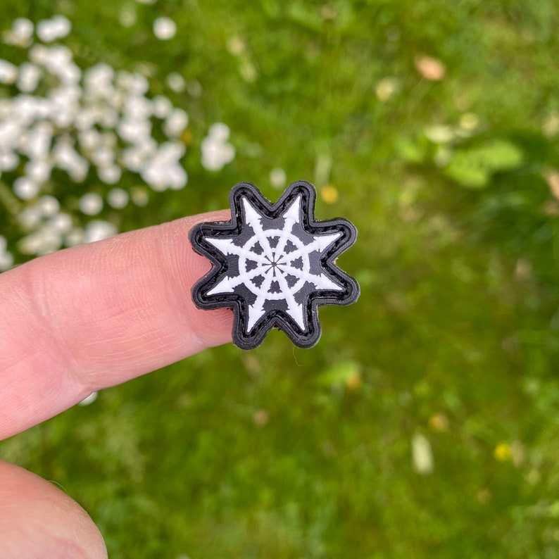 Puede incluir: Un parche negro y blanco con un dise&ntilde;o de estrella. La estrella tiene un c&iacute;rculo central con l&iacute;neas radiales. El parche est&aacute; en un dedo.