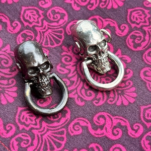 May include: Two skull-shaped pendants with large circular bails. One pendant is silver, the other is dark gray. The pendants are set against a purple background with a pink floral pattern.