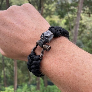 May include: Black paracord bracelet with a skull-shaped clasp. The clasp is made of metal and has a detailed design.