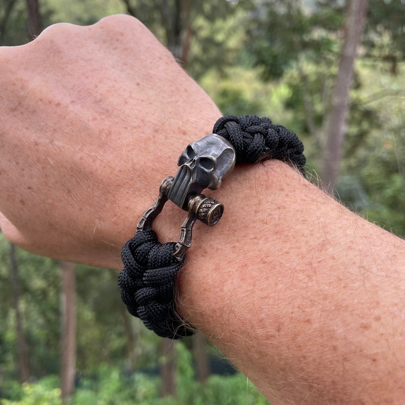 May include: Black paracord bracelet with a skull-shaped clasp. The clasp is made of metal and has a detailed design.