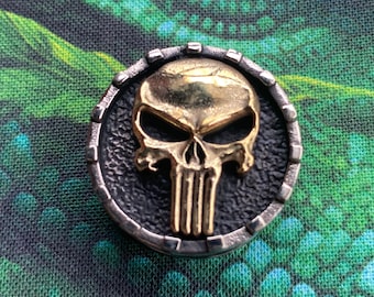 May include: A gold-colored skull with a black background, surrounded by a silver-colored ring. The skull has a menacing expression with a prominent jawline and a detailed design.