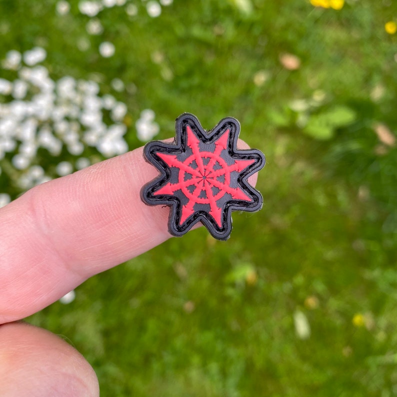 Puede incluir: Un parche en forma de estrella negro y rojo con un dise&ntilde;o circular en el centro. El parche est&aacute; hecho de tela y tiene una textura rugosa.