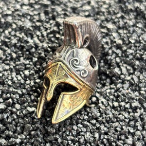 May include: A metallic Spartan helmet pendant featuring a dual-tone finish with a dark, weathered silver-coloured crest and a polished gold-coloured face guard. The helmet displays intricate embossed scrollwork details and a prominent central ridge.