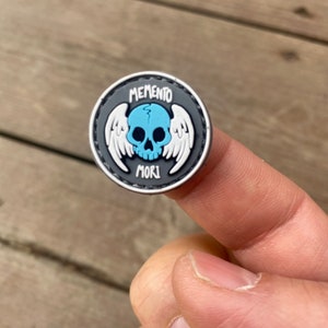 Puede incluir: Un parche blanco y negro con una calavera azul y alas. El texto "MEMENTO MORI" está escrito en el parche.