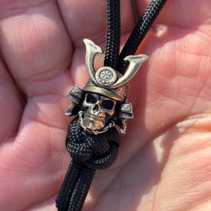 Könnte beinhalten: Ein bronzefarbener Schädelanhänger mit Samurai-Helm-Design, auf einer schwarzen Kordel. Der Helm hat dekorative Elemente und ein detailliertes Totenkopfgesicht. Die Kordel ist unterhalb des Anhängers geknotet.
