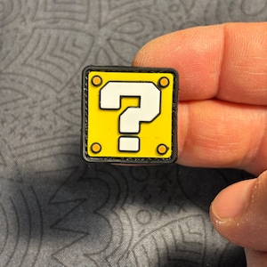 Può includere: Un ciondolo quadrato giallo con bordo nero e punto interrogativo bianco. Il ciondolo ha quattro cerchi arancioni agli angoli. Il ciondolo è tenuto dalle dita di una persona.