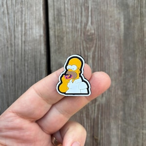 Może przedstawiać: Żółta postać z kreskówki w białej koszuli jedząca różowo-brązowy pączek. Postać jest przypinką z czarnym obramowaniem.