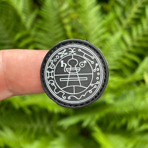 Puede incluir: Un parche blanco y negro con un diseño circular que presenta un símbolo con un círculo, líneas y una figura con cabeza y brazos. El símbolo está rodeado por un círculo con una serie de símbolos y letras.
