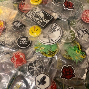 Puede incluir: Una colección de parches bordados de colores con varios diseños, incluyendo calaveras, una cara sonriente, un árbol, un kraken y un cuervo. Los parches están empaquetados en bolsas de plástico individuales.