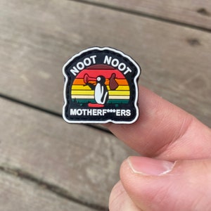 以下が含まれることがあります： 「NOOT NOOT」と「MOTHERF***ERS」の文字が入った、メガホンとバッグを持った漫画のペンギンが描かれた、黒と白の靴チャーム。夕焼けを背景にしています。