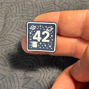 May include: A square, navy blue charm with a white border. The charm features the number "42" in white, surrounded by white stars, a UFO, and a planet. A small white towel is at the bottom. The charm is held by a person's fingers.