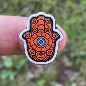 May include: A Hamsa hand charm with an orange and black design, featuring an eye symbol in the centre. The charm has a white border and is held up against a blurred green background.