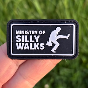 以下が含まれることがあります： 「MINISTRY OF SILLY WALKS」の文字と、おかしな歩き方をしているスーツ姿の人物のグラフィックが描かれた白黒のパッチ。パッチは黒い縁取りと、文字とグラフィック用の白い背景を持っています。