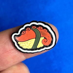 以下が含まれることがあります： オレンジ色のタコの触手デザイン、黄色いご飯、緑色の海苔巻きが特徴的な、カラフルな寿司をテーマにしたチャーム。チャームは白と黒の縁取りがあり、青色の背景に配置されています。