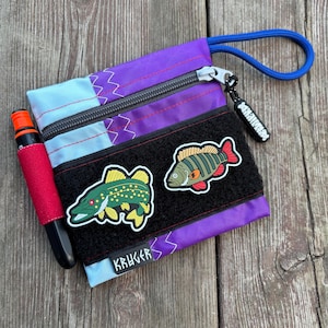 Op de afbeelding: Een paarse en blauwe ritszak met een zwarte band en twee vispatches. De vispatches zijn een groene en gele snoek en een rode en oranje baars. De tas heeft het woord "KRUGER" onderaan gedrukt.