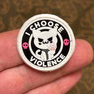 Puede incluir: Parche blanco con borde negro y el texto "I CHOOSE VIOLENCE" en un círculo. Un gato blanco con almohadillas rosas y ojos negros está dentro del círculo. Dos calaveras y huesos cruzados rosas están a cada lado del círculo.
