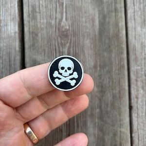 May include: A black and white enamel pin featuring a skull and crossbones design. The skull is white with black eyes and the crossbones are white.