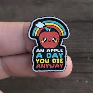 May include: A black and white cartoon apple with a rainbow above it. The apple has a happy face and is wearing a red, yellow, and green stem. The text below the apple reads "An apple a day you die anyway".