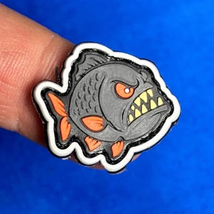 Könnte beinhalten: Ein Cartoon-Fisch-Charm mit einem bedrohlichen Ausdruck. Der Fisch ist hauptsächlich grau mit orangefarbenen Flossen und einem weißen und schwarzen Rand. Sein Maul ist geöffnet und zeigt scharfe gelbe Zähne, und er hat ein wütendes orangefarbenes Auge.