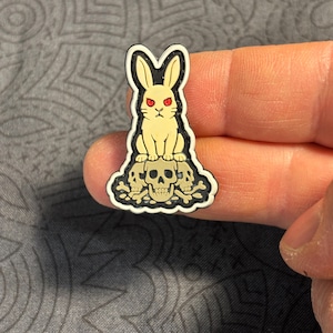May include: A cream-colored bunny with red eyes sits atop three skulls and crossbones. The bunny and skulls are outlined in black and white. This decorative pin is a unique accessory.