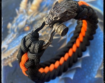 May include: A black and orange paracord bracelet with a wolf head clasp. The clasp is made of metal and has a detailed design.