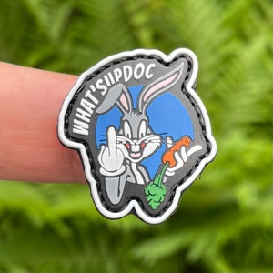 Può includere: Una toppa bianca e nera con un coniglio dei cartoni animati che tiene una carota. Il coniglio sta facendo il dito medio e il testo "WHAT'S UP DOC" è scritto intorno al coniglio.