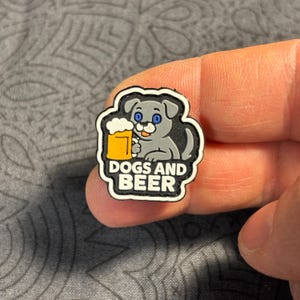 Può includere: Un piccolo ciondolo decorativo con un cane grigio dei cartoni animati con occhi blu che tiene in mano una tazza di birra. Il ciondolo ha un bordo nero e le parole "DOGS AND BEER" in bianco e giallo.