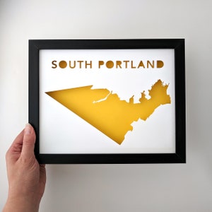 Peut inclure: Une impression encadr&eacute;e noire avec un fond blanc et une silhouette jaune des limites de la ville de South Portland, Maine. Le texte "SOUTH PORTLAND" est imprim&eacute; en noir en haut de l'impression.