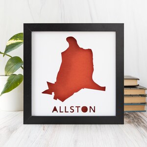 May include: A black framed print featuring a red silhouette of the Allston neighbourhood in Boston, Massachusetts. The word "Allston" is printed in red below the silhouette.