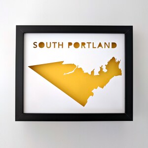 Peut inclure: Une impression encadr&eacute;e noire avec un fond blanc et une silhouette jaune des limites de la ville de South Portland, Maine. Le texte "SOUTH PORTLAND" est imprim&eacute; en noir en haut de l'impression.