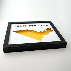 Peut inclure: Une impression encadr&eacute;e noire avec une silhouette jaune de South Portland, Maine. Le texte "SOUTH PORTLAND" est imprim&eacute; en noir en haut de l'impression.