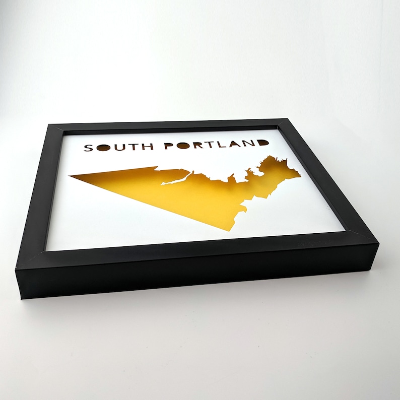 Peut inclure: Une impression encadr&eacute;e noire avec une silhouette jaune de South Portland, Maine. Le texte "SOUTH PORTLAND" est imprim&eacute; en noir en haut de l'impression.