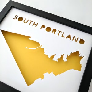 Peut inclure: Une impression encadr&eacute;e blanche avec une silhouette jaune du quartier de South Portland, dans le Maine. Le texte "SOUTH PORTLAND" est d&eacute;coup&eacute; dans le fond blanc.