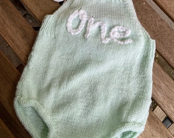 May include: A light green knitted baby onesie with the word "one" embroidered in white yarn.