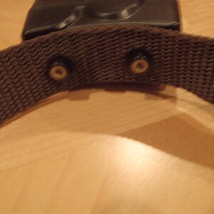 Puede incluir: Una correa de tela marr&oacute;n con dos hebillas met&aacute;licas. La correa est&aacute; unida a un objeto de pl&aacute;stico negro.