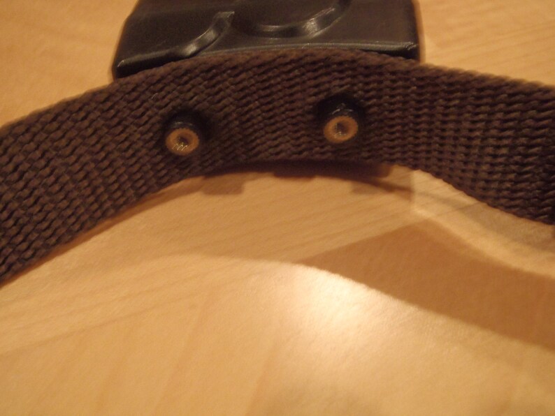 Puede incluir: Una correa de tela marr&oacute;n con dos hebillas met&aacute;licas. La correa est&aacute; unida a un objeto de pl&aacute;stico negro.