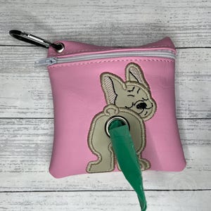 May include: Pink dog poo bag holder with a white zipper and a cute embroidered dog design. The holder has a green dispenser for the bags.