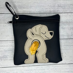 May include: Black dog waste bag dispenser with a zip closure and a silver carabiner clip. The bag features an embroidered design of a dog's rear end with a roll of yellow waste bags coming out.