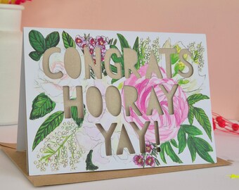 May include: A white greeting card featuring die-cut letters that spell out CONGRATS HOORAY YAY! The card is decorated with a watercolor illustration of pink and white flowers with green leaves. The text reveals a tan-colored inner layer.