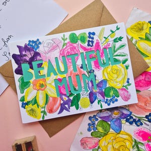 May include: A white greeting card featuring the text BEAUTIFUL MUM in bold teal letters, surrounded by a colorful watercolor illustration of various flowers including yellow roses, pink roses, purple irises, and blue forget-me-nots.