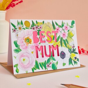 May include: A white greeting card featuring a watercolor floral illustration with pink, yellow, and green botanical elements. The words BEST MUM are die-cut into the center of the card, revealing a bright pink layer underneath the white surface.