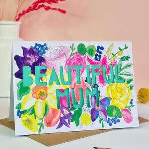 May include: A white greeting card featuring a colorful watercolor floral illustration with the die-cut text BEAUTIFUL MUM in a bright teal color. The flowers include yellow, pink, purple, and blue blossoms with green leaves.