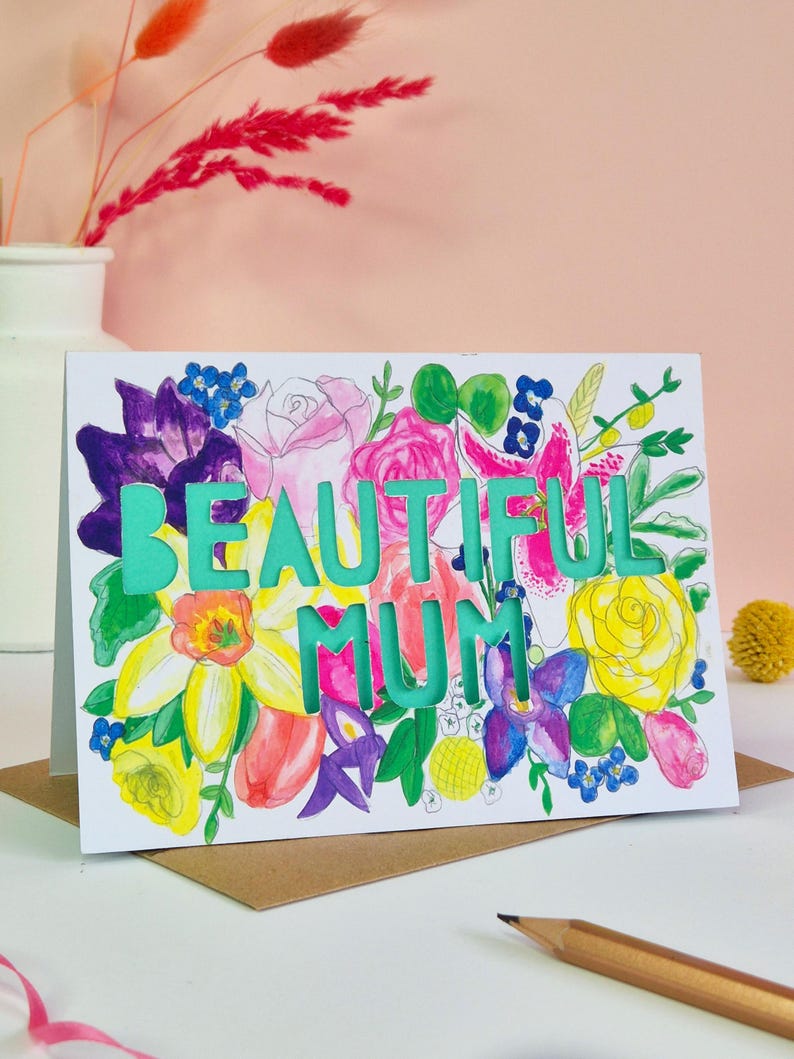 May include: A white greeting card featuring a colorful watercolor floral illustration with the die-cut text BEAUTIFUL MUM in a bright teal color. The flowers include yellow, pink, purple, and blue blossoms with green leaves.