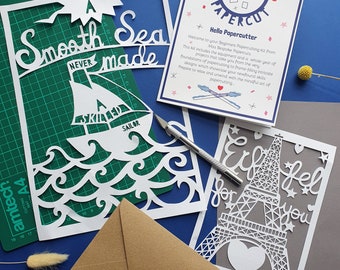 May include: Three papercutting projects, a sailboat on a blue background, the Eiffel Tower on a grey background, and a card with the text "Learn to Papercut" on a white background.