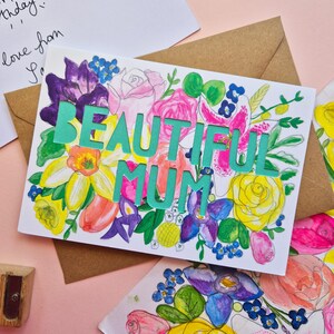 May include: A rectangular greeting card featuring a vibrant watercolor floral illustration in shades of pink, yellow, purple, and green. The text BEAUTIFUL MUM is die-cut in a bold, teal-colored font across the center of the floral design.