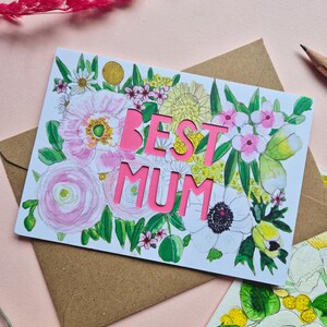 May include: A rectangular greeting card featuring a watercolor floral illustration with pink, yellow, and green botanical elements. The words BEST MUM are die-cut in bold, bright pink capital letters in the center of the card design.