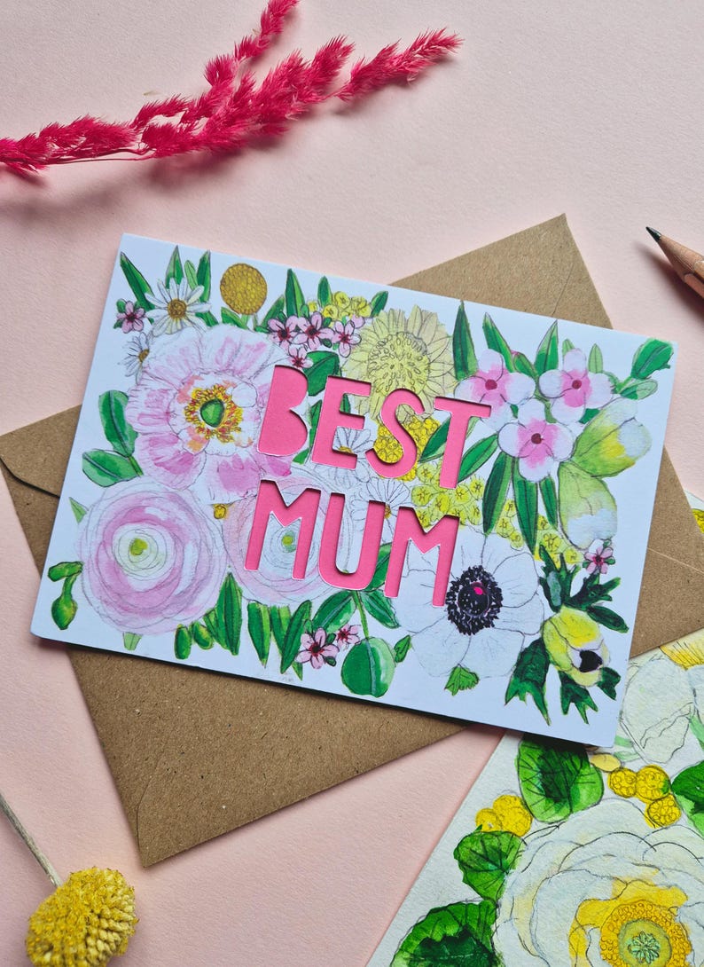 May include: A rectangular greeting card featuring a watercolor floral illustration with pink, yellow, and green botanical elements. The words BEST MUM are die-cut in bold, bright pink capital letters in the center of the card design.